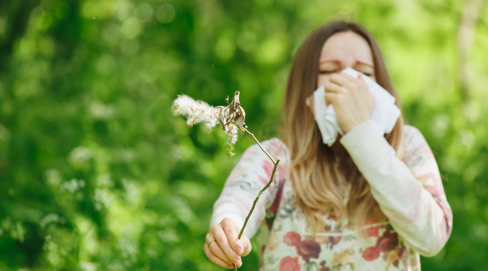 A women is outside in the garden holding a tissue to her face because she is about to sneeze. In her other hand she is holding a flower.